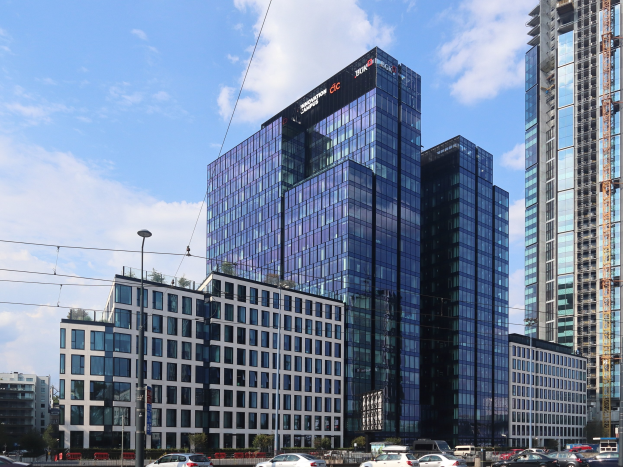 Außenansicht des Deutschen Bank-Hauptsitzes in Frankfurt, Deutschland, mit einem mehrstöckigen Gebäude mit zahlreichen Fenstern, umgeben von städtischer Infrastruktur wie Straßenlaternen, Fahrzeugen, Grünflächen und einem bewölkten Himmel.