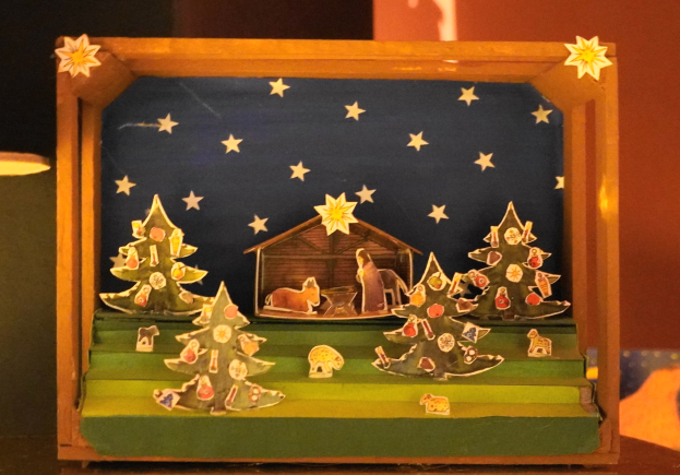 Eine hölzerne Krippe auf einem Tisch, mit einer Lampe links und einer dekorierten Schachtel mit Weihnachtsbäumen, Tieren und einem Haus im Hintergrund.