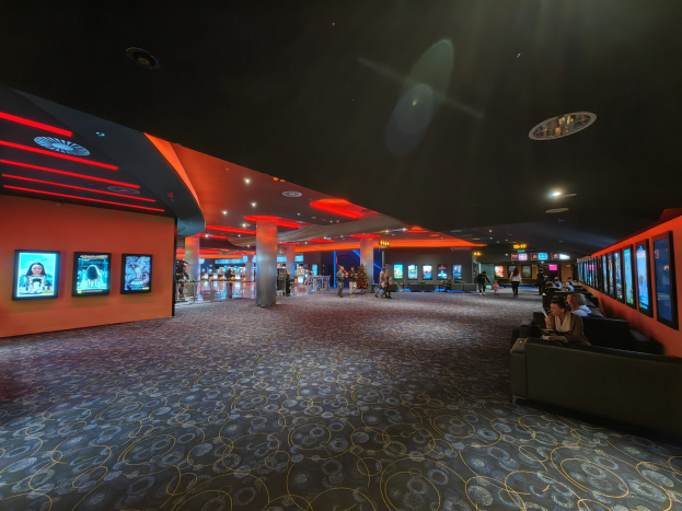 Großer Raum mit Menschen auf Sofas sitzend, die an der Wand montierte Fernseher beobachten, umgeben von Säulen und Deckenleuchten, mit sichtbarem Boden.