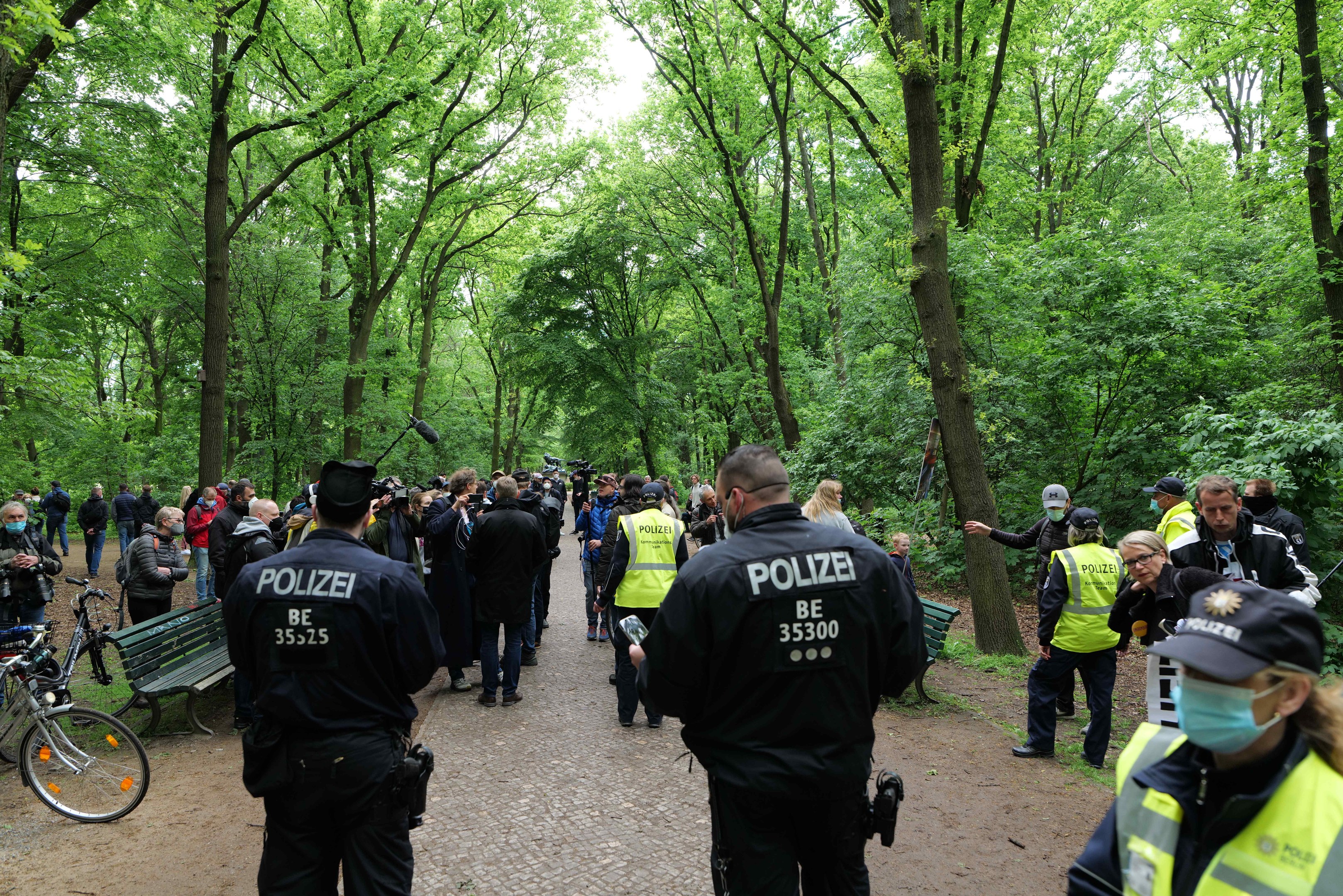 Polizisten vor einer Menge bei einer Outdoor-Veranstaltung, mit Fahrrädern und einer Bank im Vordergrund, Bäumen und Himmel im Hintergrund.
