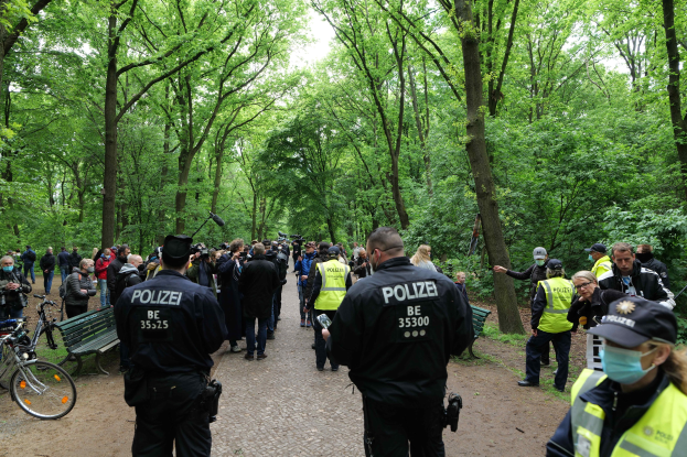 Polizisten vor einer Menge bei einer Outdoor-Veranstaltung, mit Fahrrädern und einer Bank im Vordergrund, Bäumen und Himmel im Hintergrund.