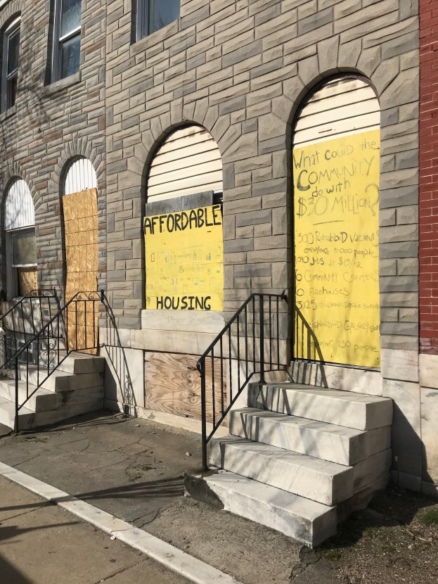 Ein verbarrikadiertes Gebäude mit einer Tafel, die "Bezahlbarer Wohnraum" anzeigt, eine Treppe mit Geländer, die zum Eingang führt, und eine Straße unten.