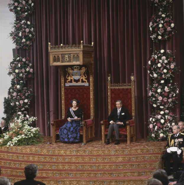 Königin Elizabeth II und Prinz Philip, Herzog von Edinburgh, sitzen vor einem Publikum, wobei die Königin ein blaues Kleid trägt und ein Buch hält, und der Herzog eine schwarze Jacke, ein weißes Hemd und schwarze Hosen trägt, vor einem maronenfarbenen Vorhang mit Blumenbouquets auf beiden Seiten.