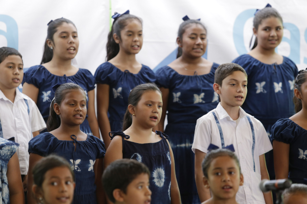 Eine Gruppe von Kindern vor einem Mikrofon mit einem Schulchor-Banner im Hintergrund, die zu singen scheinen.