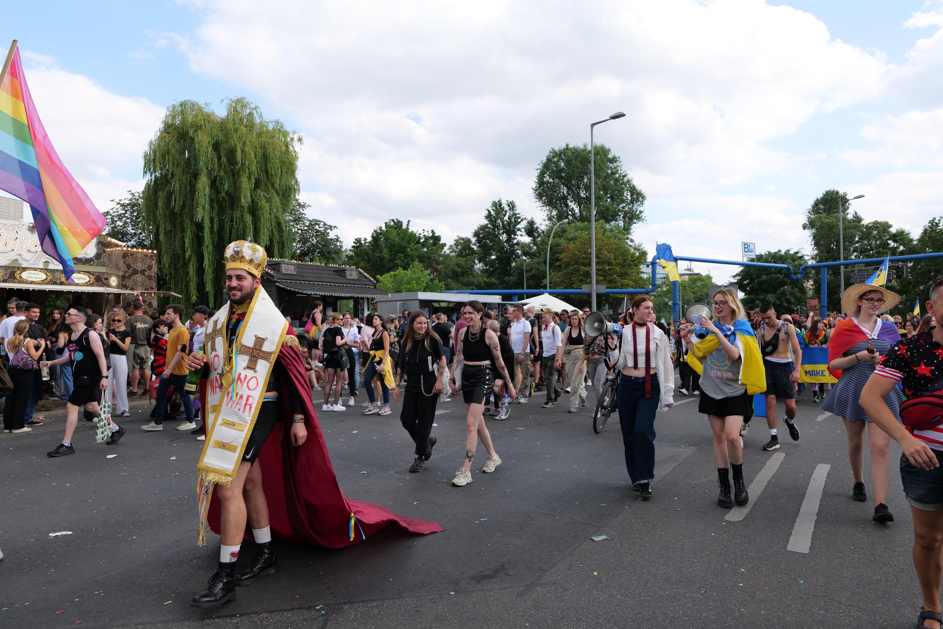 Eine Gruppe von Menschen marschiert auf der Pride Parade 2018 mit einer Regenbogenflagge und Musikinstrumenten, mit Laternenmasten, Bäumen und Hütten im Hintergrund.