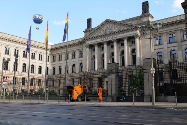 Großes beige Gebäude mit zahlreichen Fenstern, der Deutsche Bundestag in Berlin, flankiert von Straßenmöbeln, Fahrzeugen, Fußgängern, Flaggen und Bäumen unter einem bewölkten Himmel.