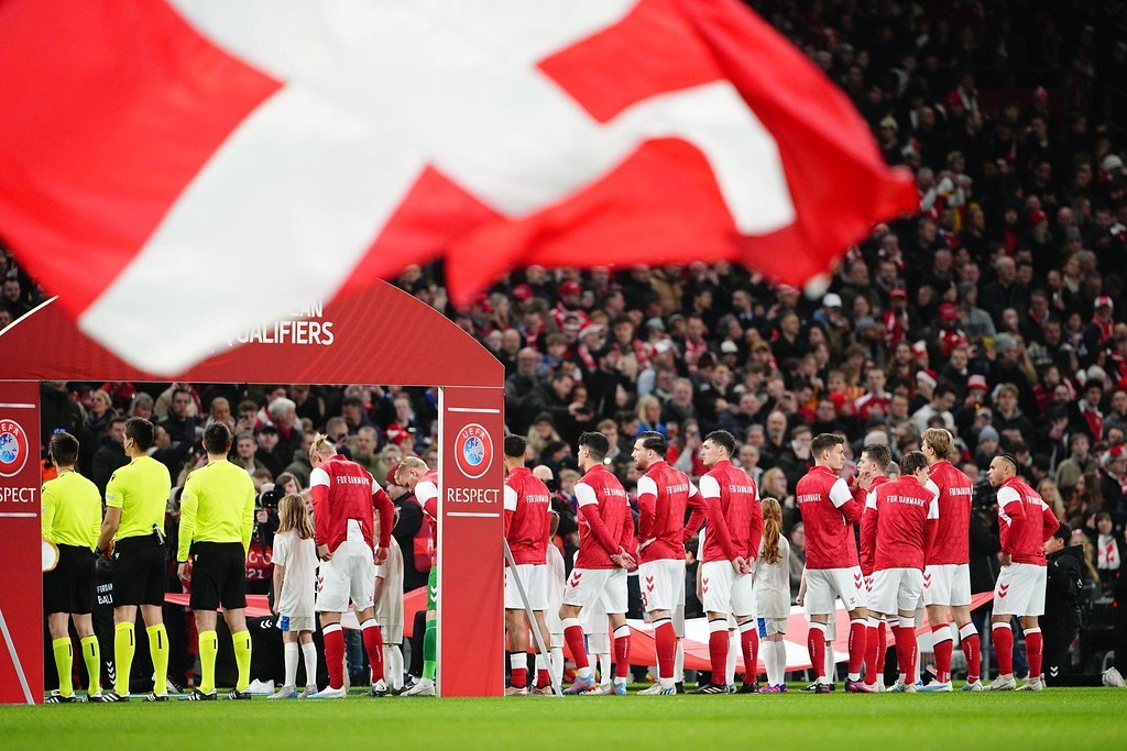 Eine Gruppe von Menschen auf einem Fußballfeld mit einer roten und weißen Flagge im Vordergrund, ein Bogen mit der Aufschrift "Bayern Munich vs Bayern Munich Betting Tips & Preview" im Hintergrund und eine große Menge im Stadion.