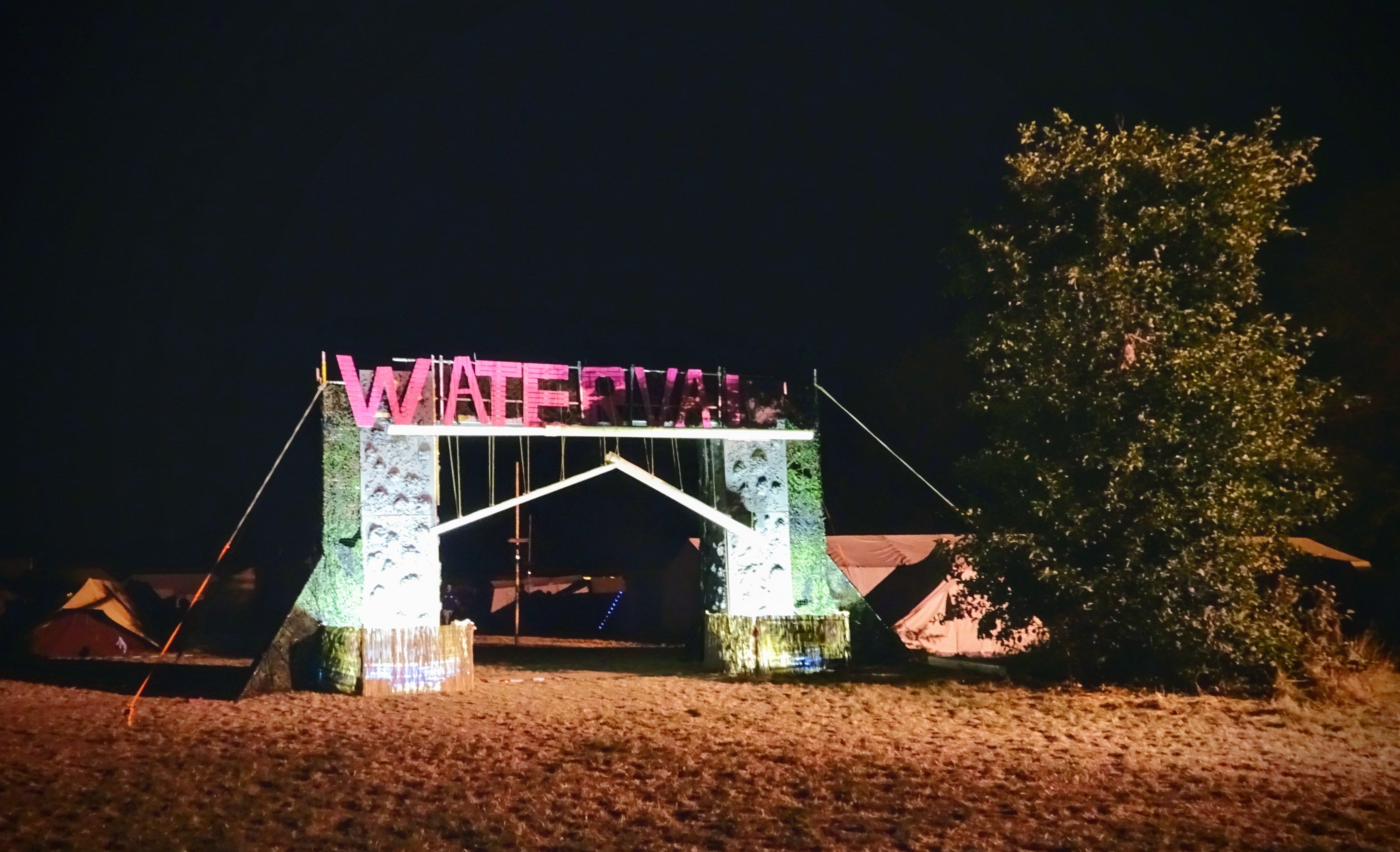 Bühne für ein nächtliches Musikfestival mit einem Bogen mit Text, einem Baum auf der rechten Seite, Zelten im Hintergrund und einem dunklen Himmel.