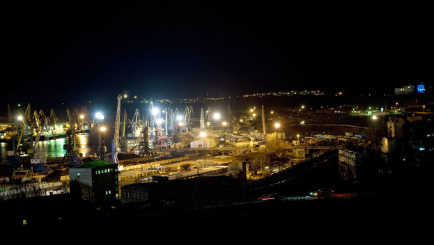 Eine nächtliche Stadtansicht mit beleuchteten Gebäuden, fahrenden Fahrzeugen auf der Straße und Spiegelungen im Wasser vor einem dunklen Himmel.