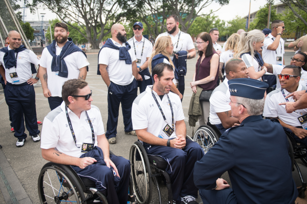 Eine Gruppe von Menschen im Rollstuhl, die vor einer Kulisse aus Bäumen, Pfählen, Gebäuden und einem klaren blauen Himmel miteinander sprechen, wobei einige Mützen und Brillen tragen, was darauf hindeutet, dass sie an den Paralympischen Spielen teilnehmen.