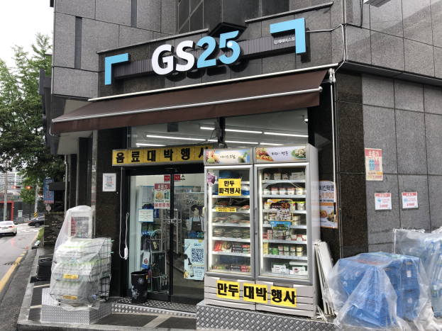 Ein Gebäude mit einem "GS25"-Schild, umgeben von Fahrzeugen, Bäumen und einem klaren blauen Himmel, das einen koreanischen Lebensmittelmarkt mit sichtbaren Kühlgeräten beherbergt, die mit Lebensmitteln gefüllt sind.