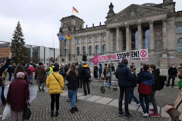 Eine Gruppe von Menschen demonstriert vor einem Regierungsgebäude mit Fenstern, Säulen und Bögen, hält Schilder und Fahnen, mit einem Baum im Vordergrund und einem bewölkten Himmel im Hintergrund.