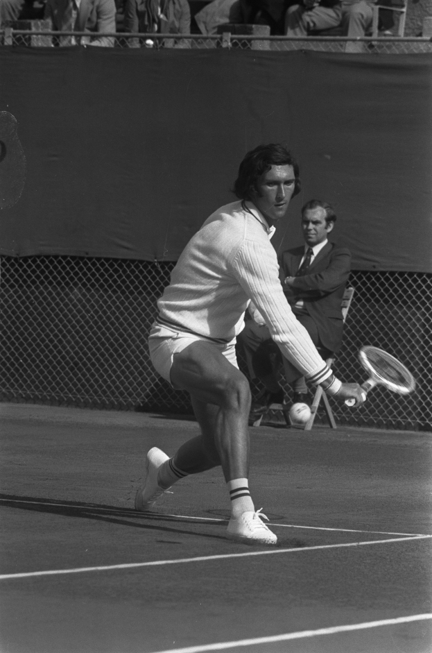 Ein professioneller Tennisspieler schlägt mit einem Schläger einen Ball während eines Spiels, mit Zuschauern und Maschendrahtzaun im Hintergrund.