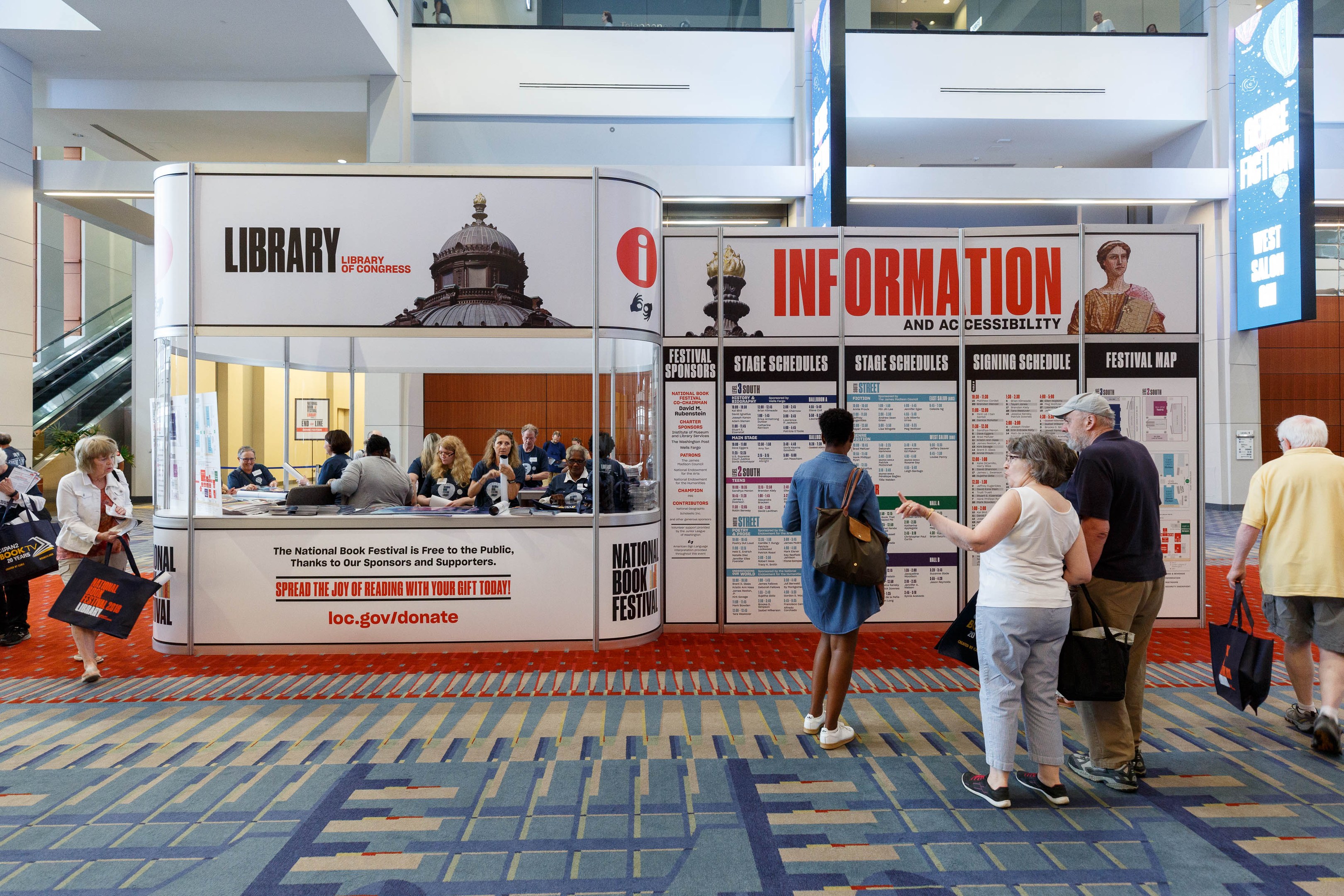 Gruppe von Menschen vor einem Stand auf der Nationalen Buchmesse versammelt, einige sitzen auf Stühlen und andere stehen, mit Schildern, Anzeigebildschirm, Treppe, Geländer und Deckenbeleuchtung.
