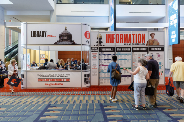 Gruppe von Menschen vor einem Stand auf der Nationalen Buchmesse versammelt, einige sitzen auf Stühlen und andere stehen, mit Schildern, Anzeigebildschirm, Treppe, Geländer und Deckenbeleuchtung.