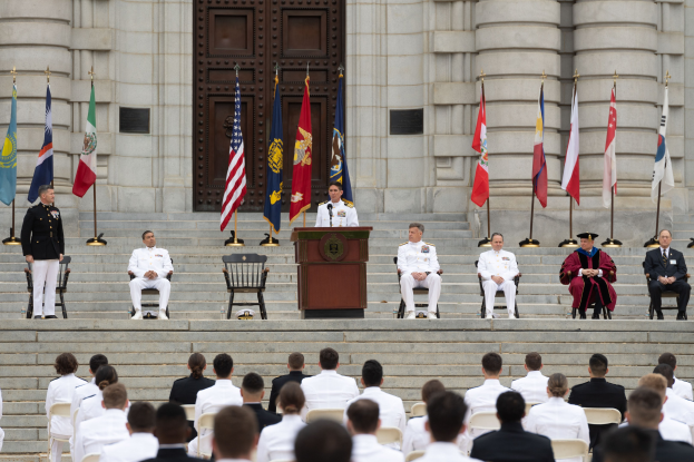 Gruppe von Menschen in weißen Uniformen auf den Stufen eines Gebäudes mit einem Podium, das von Fahnen flankiert ist, vor einem großen Gebäude mit einer Tür während einer Ehrungszeremonie.