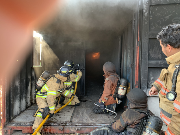 Eine Gruppe von Feuerwehrleuten auf der Ladefläche eines Trucks, mit Helmen und Zylindern, einer Person mit einer Rohrleitung, während Rauch aus dem Truck aufsteigt.