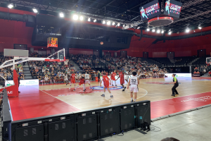 Eine Gruppe von Menschen, die Basketball in einer großen Arena spielt, mit Zuschauern im Hintergrund, einem Basketballkorb links, Deckenlampen, einem Bildschirm und schwarzen Objekten unten.