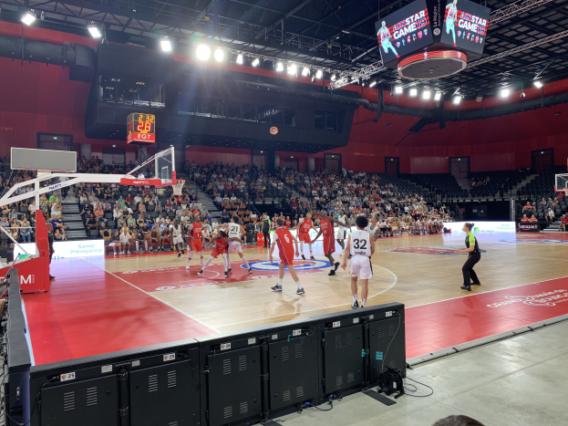 Eine Gruppe von Menschen, die Basketball in einer großen Arena spielt, mit Zuschauern im Hintergrund, einem Basketballkorb links, Deckenlampen, einem Bildschirm und schwarzen Objekten unten.