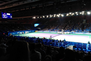 Tennis-Match in einer großen Arena mit Zuschauern auf Stühlen, ein Netz auf dem Platz, einige stehende Personen und eine Anzeigetafel mit Text links sowie Stadionbeleuchtung von oben.