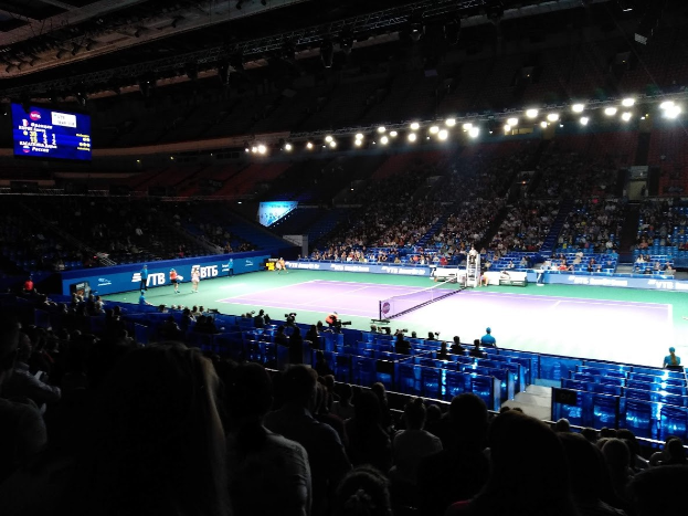 Tennis-Match in einer großen Arena mit Zuschauern auf Stühlen, ein Netz auf dem Platz, einige stehende Personen und eine Anzeigetafel mit Text links sowie Stadionbeleuchtung von oben.