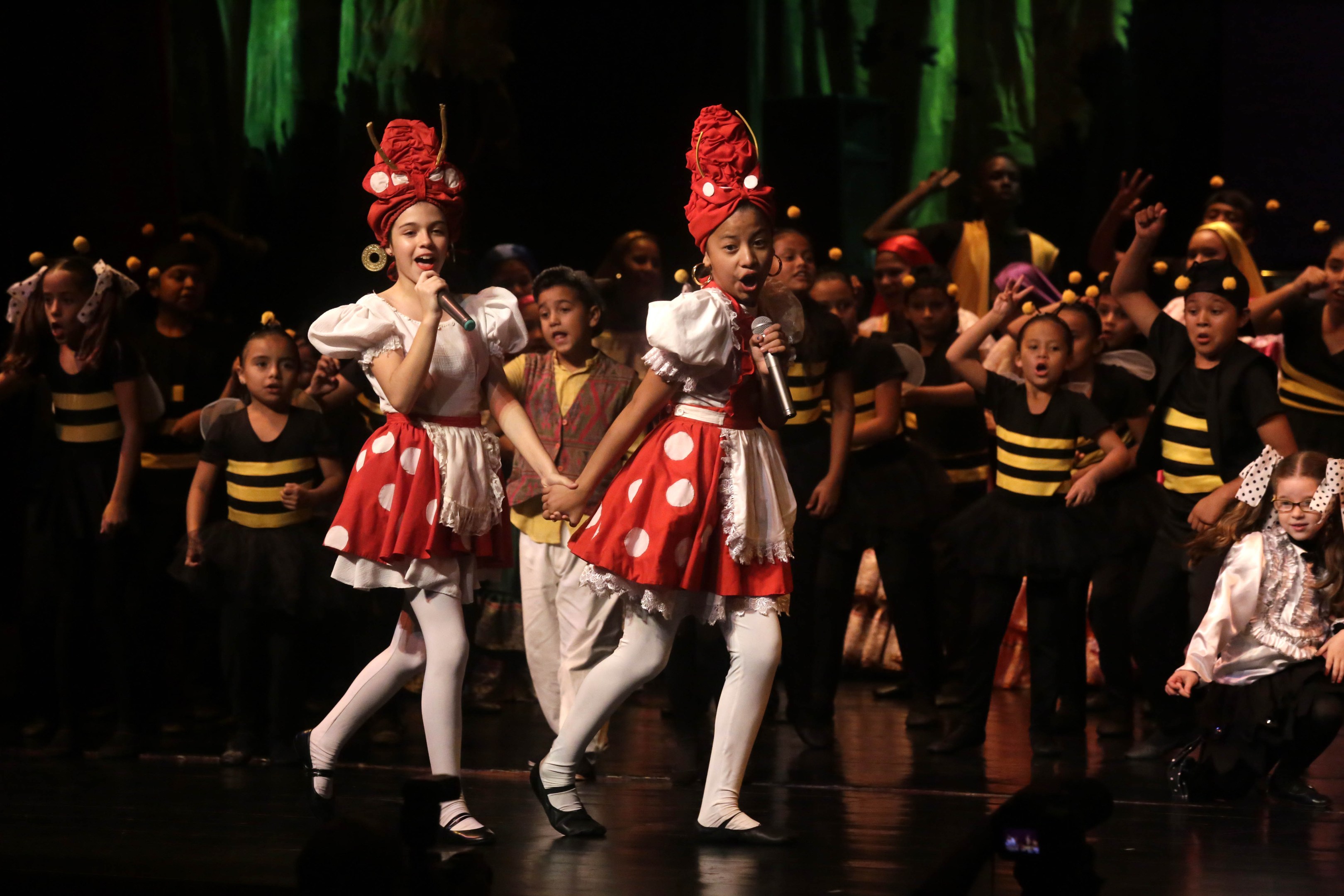 Kinder in roter und weißer Tracht auf einer Bühne tanzend, zwei mit Mikrofonen, vor dunklem Hintergrund und Zuschauern.