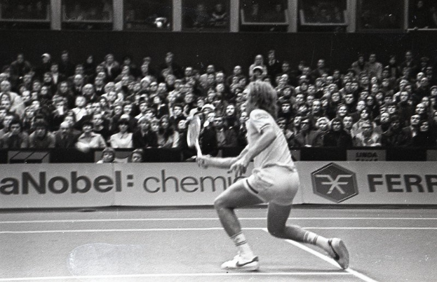 Schwarzes und weißes Bild einer Frau, die Tennis auf einem Platz mit Zuschauern in den Rängen und einem Banner im Hintergrund spielt.