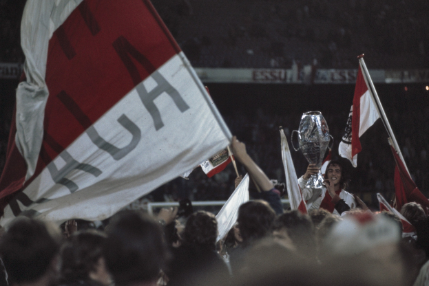 Eine Gruppe von Menschen mit Fahnen und einem Pokal vor einem jubelnden Stadionpublikum, wobei die zentrale Figur den Pokal stolz präsentiert.