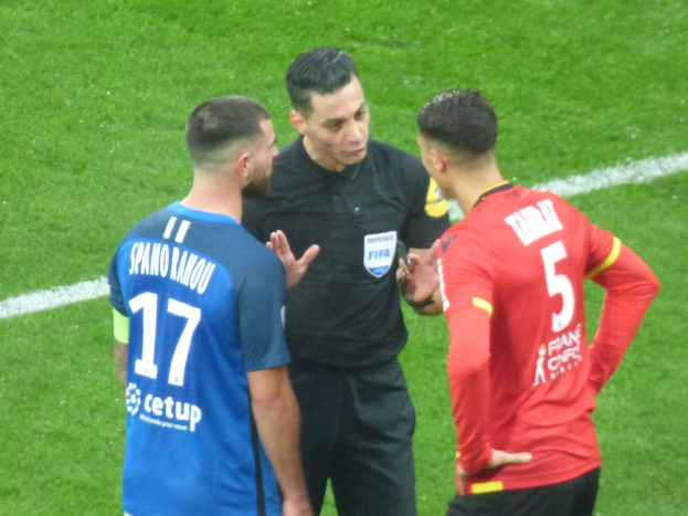 Schiedsrichter in schwarzem Hemd und Hose, der mit zwei Männern in blauer und roter Kleidung spricht, mit Text, alle tragen Brillen, auf einem Fußballfeld.