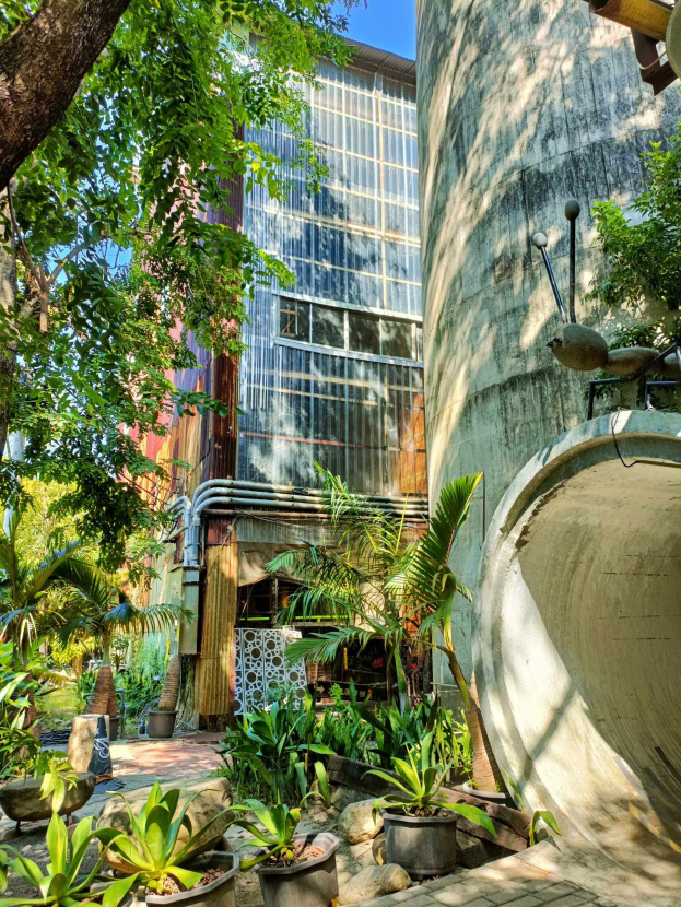 Ein Innenhof mit Topfpflanzen und Bäumen vor einem modernen umweltfreundlichen Hotel, umgeben von üppiger Vegetation unter einem klaren blauen Himmel.