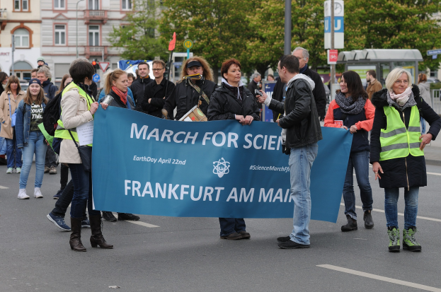 Gruppe von Menschen marschiert die Straße entlang und hält ein "March for Science Frankfurt am Main"-Schild mit Bäumen, Pfählen und Gebäuden im Hintergrund unter einem klaren blauen Himmel.