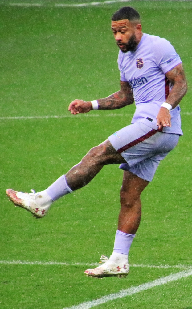 Ein Mann in einem lila Hemd und Fußballstiefeln kickt einen Fußball auf einem grünen Feld.