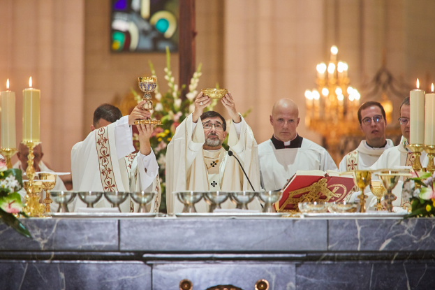 Eine Gruppe von Priestern steht vor einem Altar, jeder hält einen Kelch in der Hand, mit Gläsern, einem Mikrofon, einem Buch und anderen Gegenständen auf dem zentralen Tisch, einem Blumenstrauß links und einem Kerzenständer rechts, vor einem Hintergrund mit einem Bleiglasfenster.
