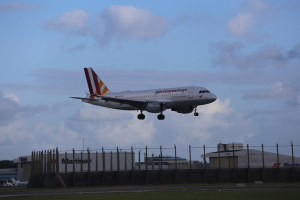 Germanwings Airbus A320-200 beim Start vom Frankfurt Airport mit Gras darunter, einem Zaun im Vordergrund, Gebäuden und Bäumen im Hintergrund und einer bewölkten Himmel.