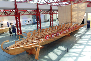 Ein großes hölzernes Wikingerschiff in einem Museum ausgestellt, umgeben von Besuchern, Metallstangen, einem Zaun, einer Fahne, einer Wand und einer Decke mit Lampen.