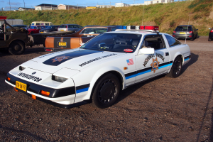 Ein Polizeiwagen auf einem Parkplatz neben anderen Autos geparkt, umgeben von Gras, Pfosten, Drähten, Gebäuden und einem klaren blauen Himmel.