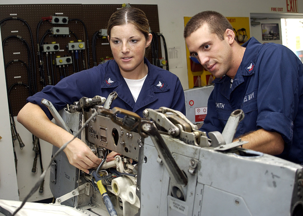 Zwei Fabrikarbeiter in blauen Uniformen bedienen Maschinen, mit industrieller Ausrüstung und einer Wandtafel im Hintergrund.