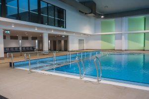 Großer Hallenbad in einem großen Gebäude mit gefliestem Boden, Metallstangen, Wänden, Säulen, Türen, Deckenleuchten und anderen baulichen Elementen.