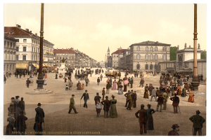 Ein altes Foto einer belebten Münchner Stadtstraße aus den frühen 1900er Jahren, das Fußgänger, einige mit Schirmen, einen Pferdewagen im Vordergrund, Gebäude, Bäume und Pfosten im Hintergrund zeigt, mit Text am unteren Bildrand.