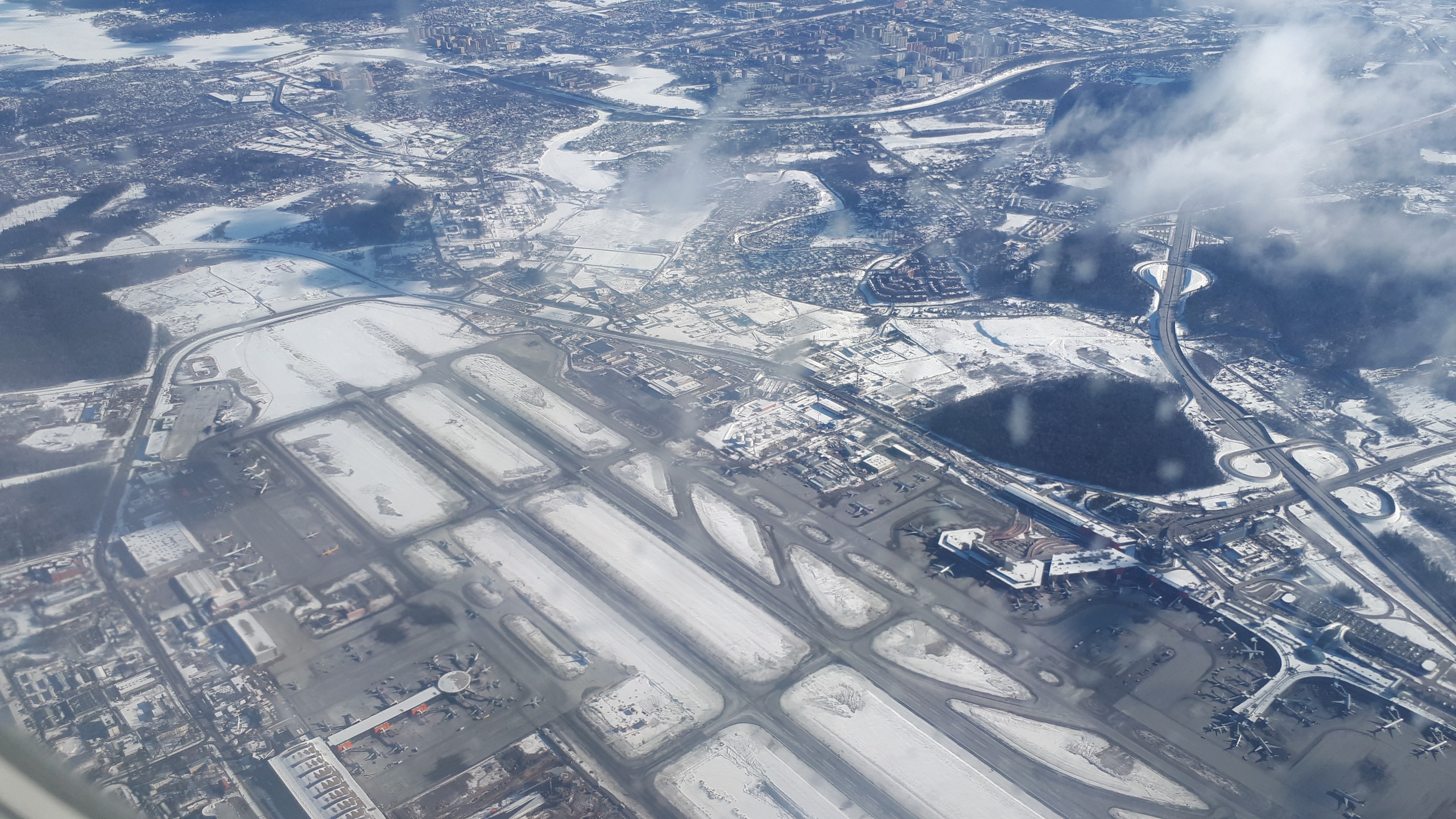 Luftaufnahme eines Flughafens mit schneebedeckter Landschaft, Gebäuden, Straßen, Brücken, Wasser, Bäumen und Wolken am Himmel.