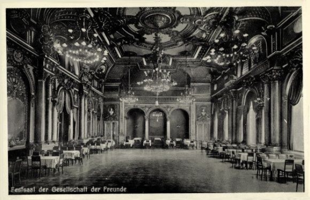 Ein altes Schwarz-Weiß-Foto eines großen Raumes mit Tischen, Stühlen, Säulen, Bögen und Kronleuchtern an der Decke, mit Text unten, der "Festival der Gesellschaft der Freunde, Berlin" lautet.