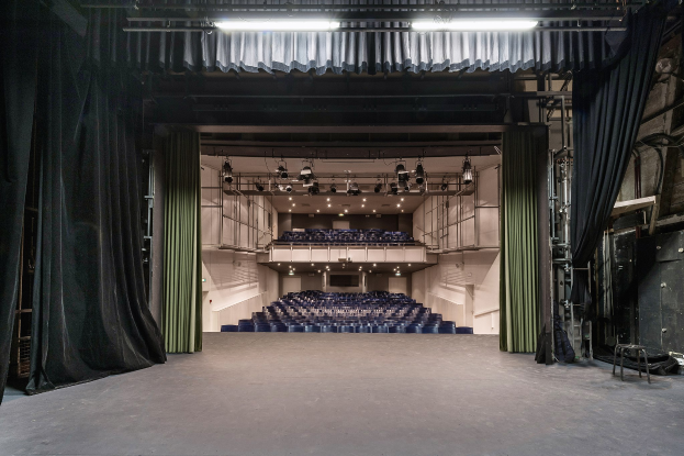 Großer Zuschauerraum mit schwarzen Vorhängen, einer Bühne, Deckenbeleuchtung, Stühlen auf dem Boden und Gegenständen auf der rechten Seite, was auf eine Veranstaltungslocation hinweist.