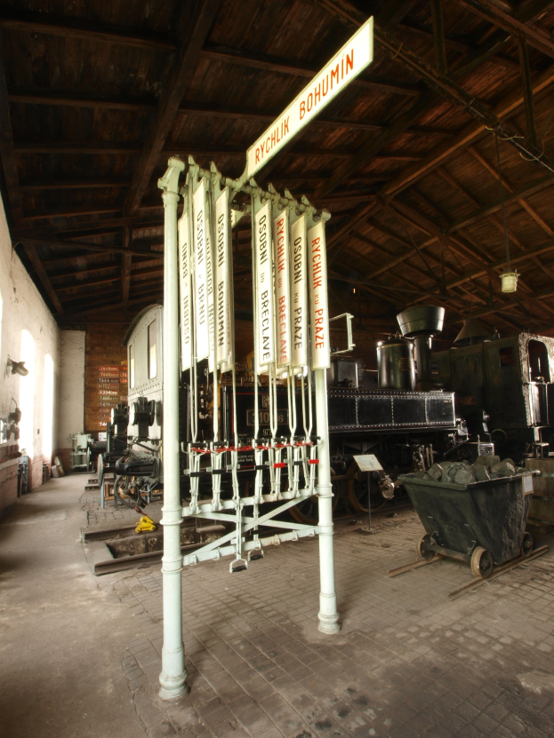 Ein Bahnsteig mit einem Zug auf den Schienen, einem Rollstuhl auf dem Boden, einem Signalkasten in der Mitte, einem Pfosten mit einer Tafel, einer Wand auf der linken Seite und einer Decke darüber.