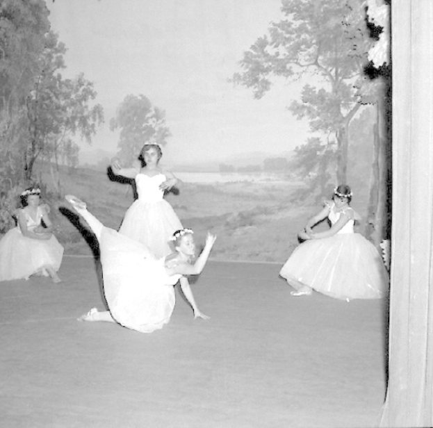 Vier junge Balletttänzerinnen in weißen Kleidern tanzen auf einer Bühne, mit einer gemalten Kulisse aus Bäumen und Himmel, in Schwarz-Weiß.