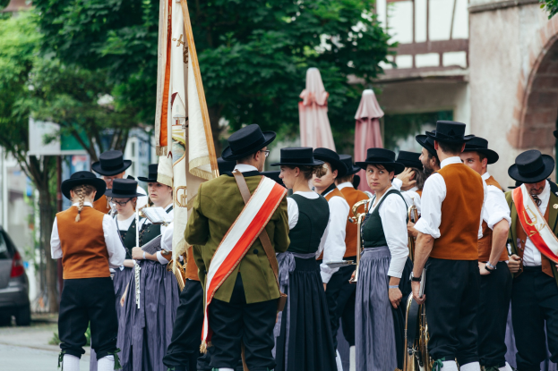 Eine Gruppe von Menschen in traditioneller bayrischer Tracht marschiert auf einer Straße, einige tragen Musikinstrumente und Fahnen, im Hintergrund sind Bäume, Gebäude und ein Auto zu sehen.