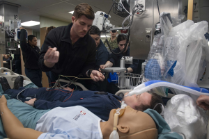 Ein Mann liegt auf einem Krankenhausbett neben einer medizinischen Puppe, während mehrere Menschen beobachten, mit medizinischer Ausrüstung und Deckenbeleuchtung in einer medizinischen Einrichtung.