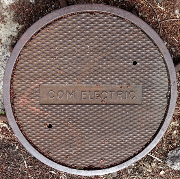 Abdeckplatte mit "com electric" Text auf dem Boden, umgeben von Steinen und Zweigen.