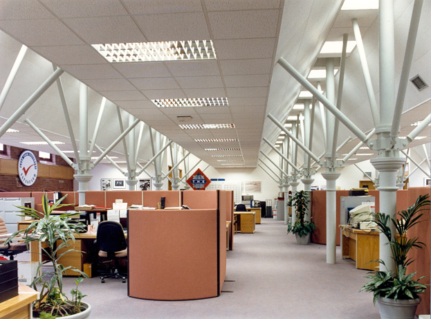 Ein geräumiges, leeres Büro mit Reihen von Schreibtischen und Stühlen, Topfpflanzen, tragenden Säulen, einer Wanduhr und Deckenleuchten in ordentlicher Anordnung.