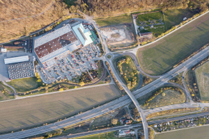Luftaufnahme eines großen Gebäudes auf einem Feld, umgeben von Straßen, Fahrzeugen, Bäumen und Gras, mit einem Logo, das darauf hinweist, dass es sich wahrscheinlich um die deutsche Automobil-Zentrale handelt.