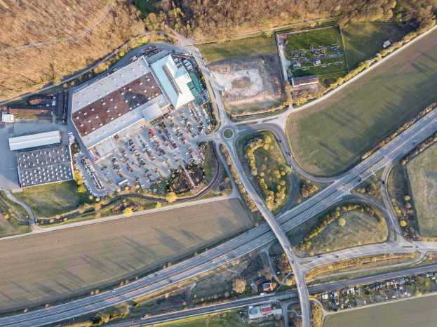Luftaufnahme eines großen Gebäudes auf einem Feld, umgeben von Straßen, Fahrzeugen, Bäumen und Gras, mit einem Logo, das darauf hinweist, dass es sich wahrscheinlich um die deutsche Automobil-Zentrale handelt.